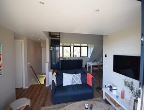 Ein modernes Wohnzimmer mit einem dunkelblauen Sofa, einem Flachbildfernseher und einer durch eine offene Tür sichtbaren Küchenzeile.