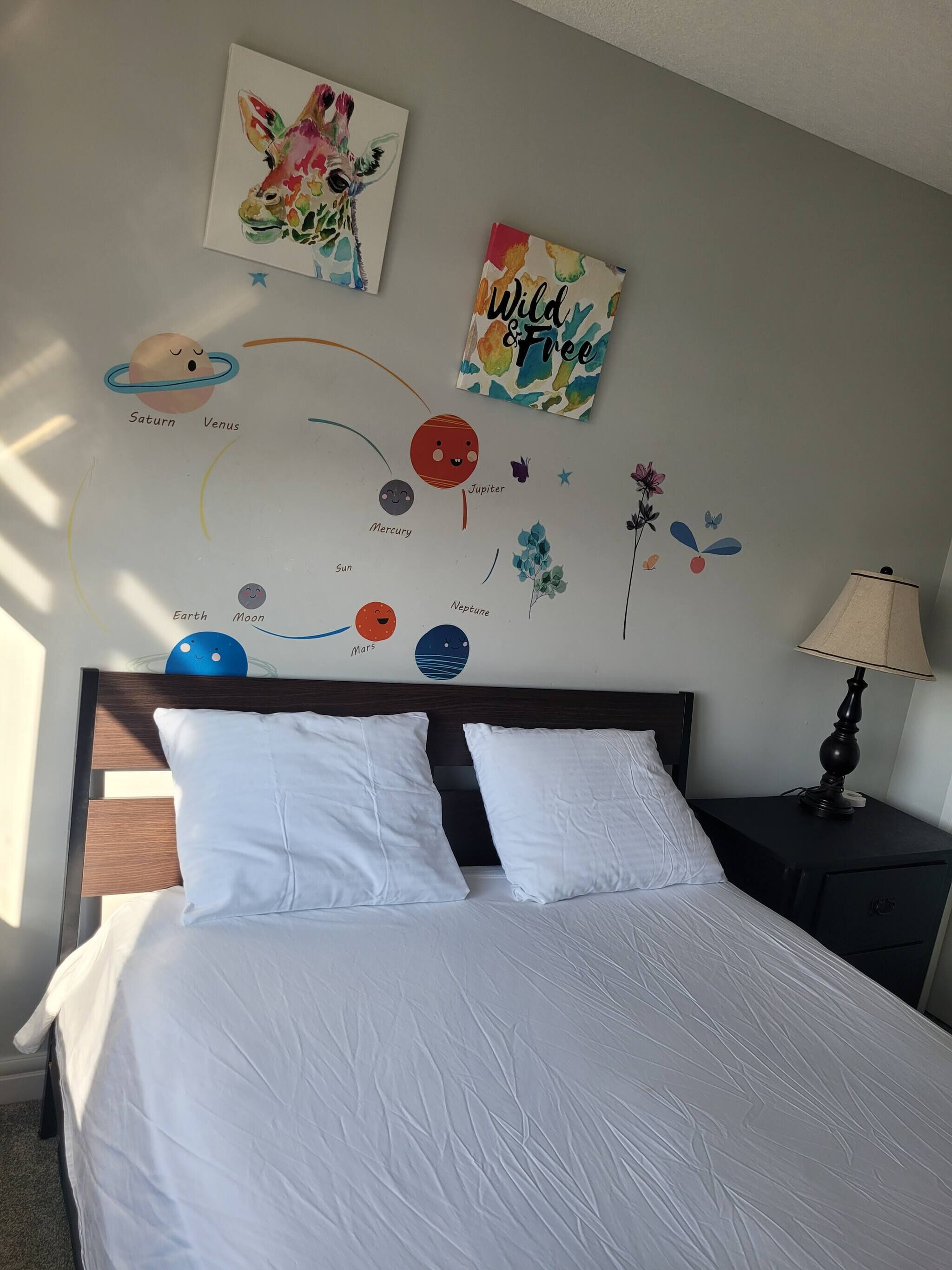 Une chambre à coucher comprenant un lit, des oreillers, une table de chevet, une lampe et des œuvres d’art mural représentant des planètes et des fleurs.