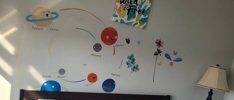 Une chambre à coucher comprenant un lit, des oreillers, une table de chevet, une lampe et des œuvres d’art mural représentant des planètes et des fleurs.