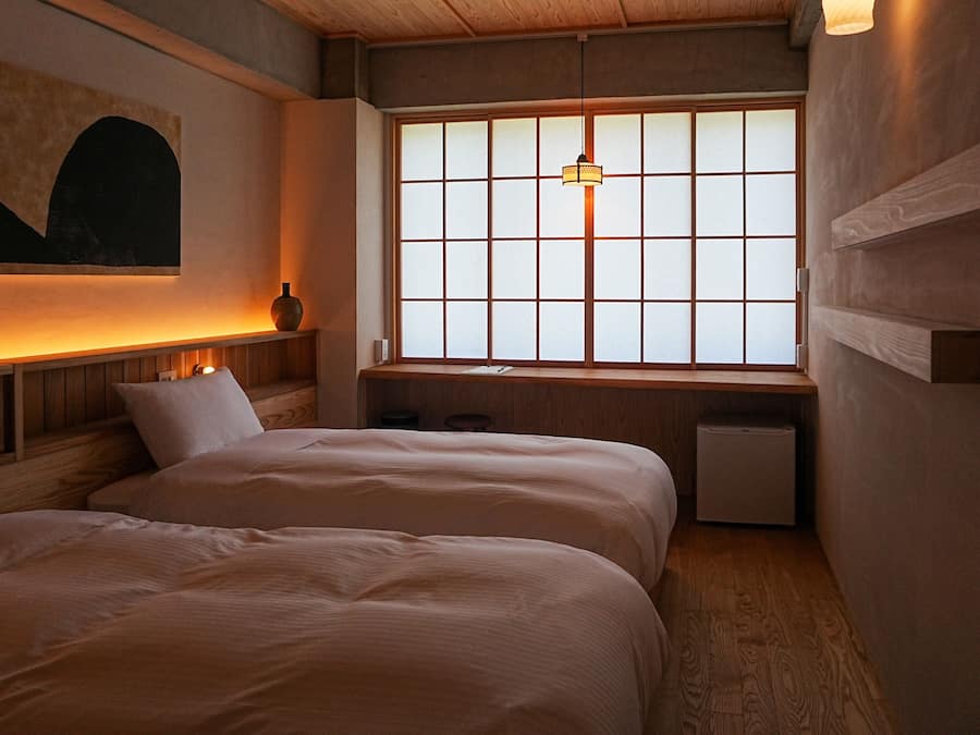 A room with two beds, a wooden ceiling, a window with a grid pattern, and a hanging light fixture.