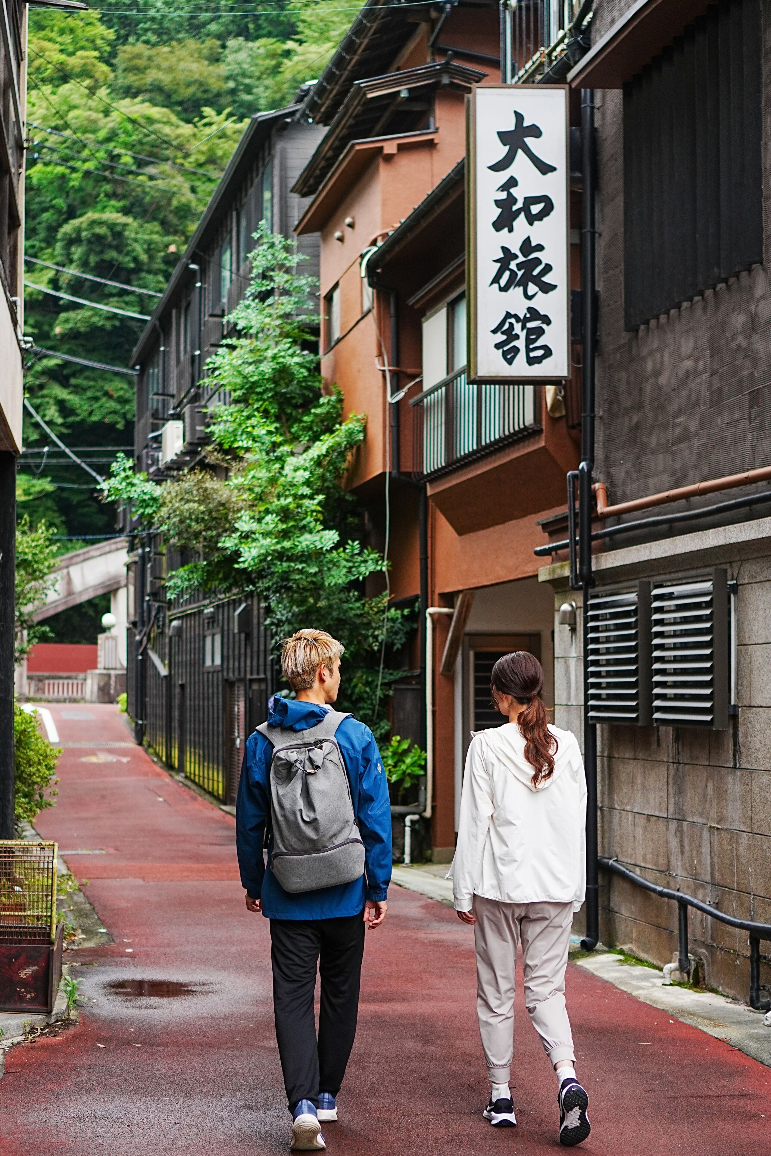 Deux personnes qui marchent dans une ruelle étroite entre deux bâtiments, avec une enseigne en japonais.
