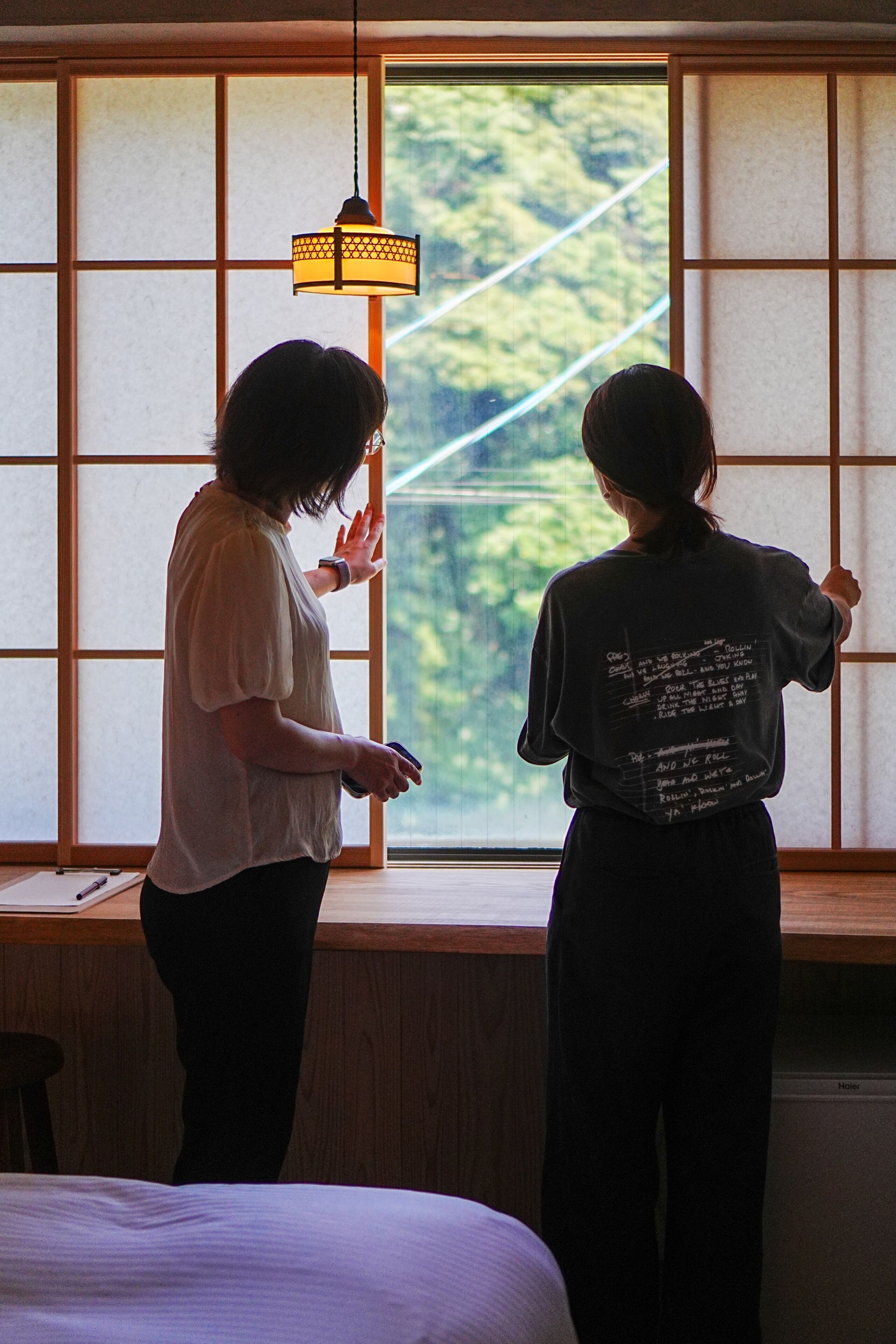Two individuals standing by a window with a view of greenery, a lamp, and a wooden desk.