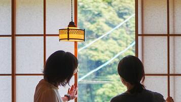 Two individuals standing by a window with a view of greenery, a lamp, and a wooden desk.
