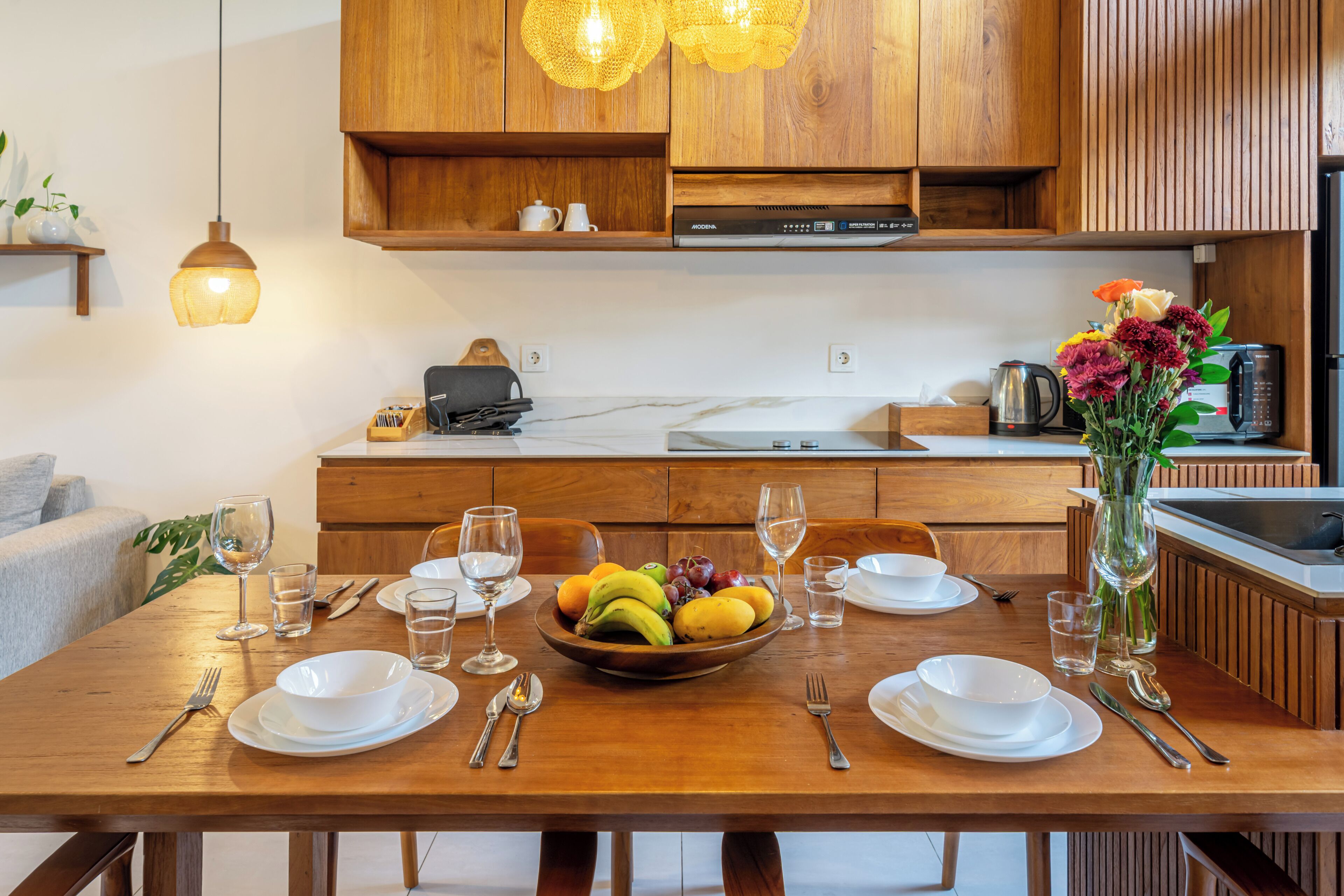 A wooden dining table with a fruit bowl, white plates, and cutlery, set in a modern kitchen with wooden cabinets and pendant lighting.