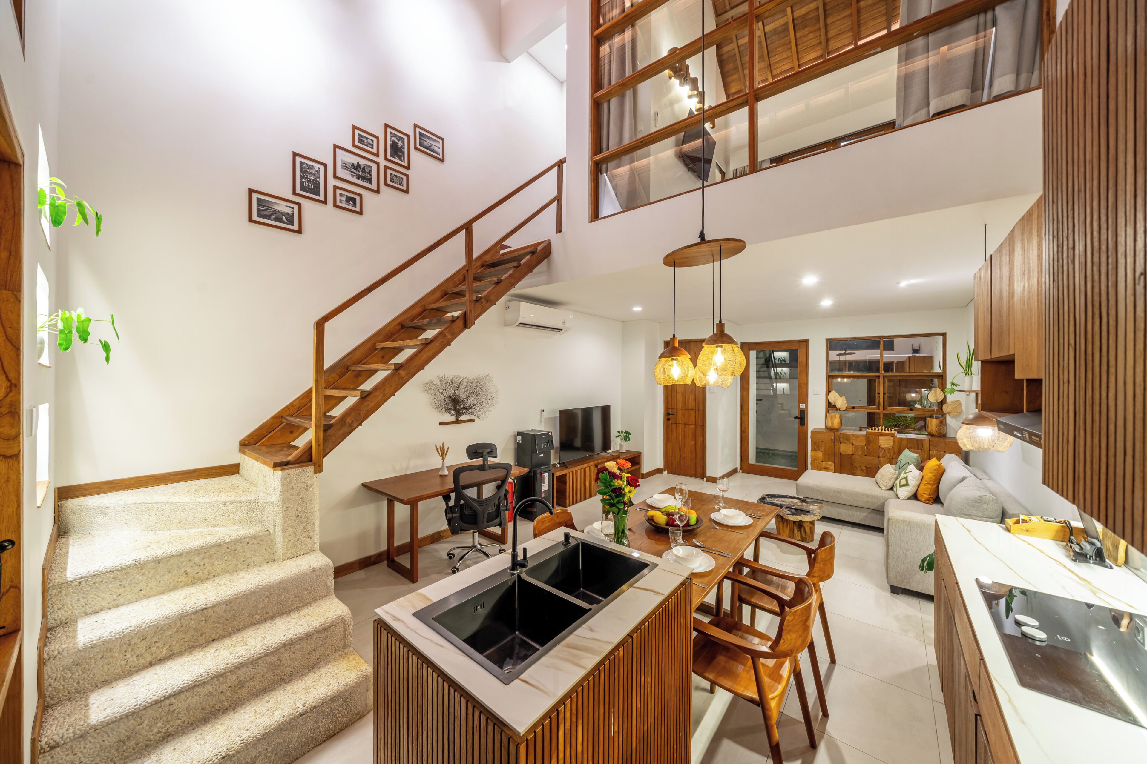 A modern kitchen with a built-in stove, a dining area with a wooden table and chairs, a staircase, and a living area with a sofa and a TV.