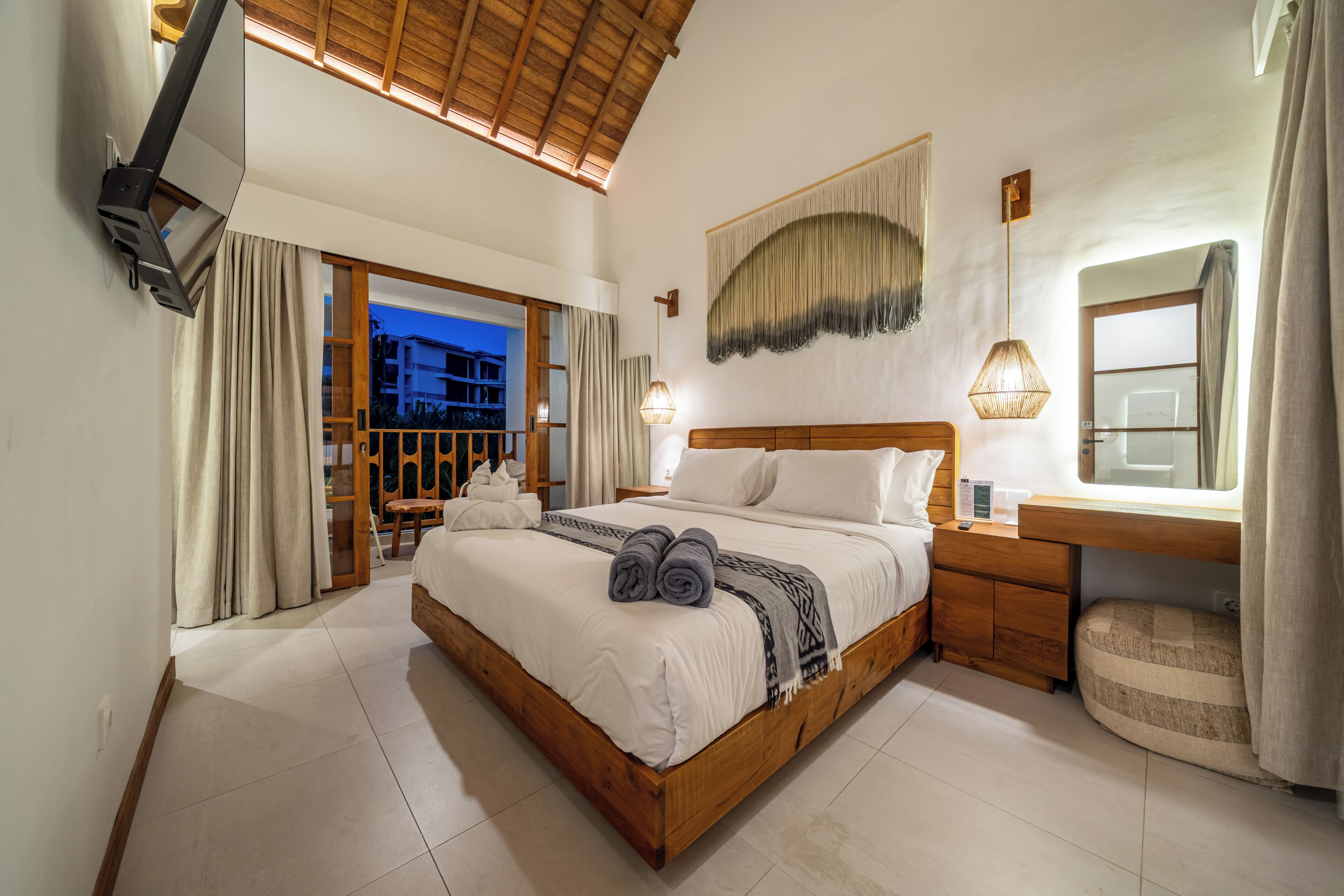 A spacious bedroom with a large bed, a wooden headboard, a bedside table with a lamp, a large mirror, and a view of the outside balcony.