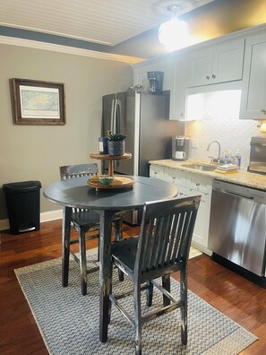 A kitchen with a dining table, chairs, a refrigerator, a coffee maker, and a trash can. - Peaceful River View Condo (Townsend)