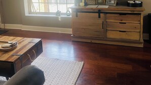 Una sala de estar con un aparador de madera, un televisor de pantalla plana, una ventana con persianas y piso de madera.
