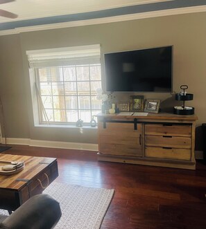 A living room with a wooden cabinet, a flat-screen TV, a window with blinds, and a wooden floor. - Peaceful River View Condo (Townsend)