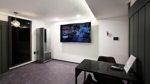 Habitación de hotel moderna con un televisor de pantalla plana grande montado en la pared, un refrigerador empotrado, una mesa de comedor con sillas y una lámpara colgante.