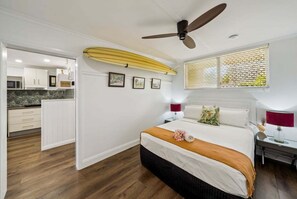 Une chambre à coucher avec un lit, un ventilateur de plafond, une fenêtre avec des stores et une vue sur la cuisine.