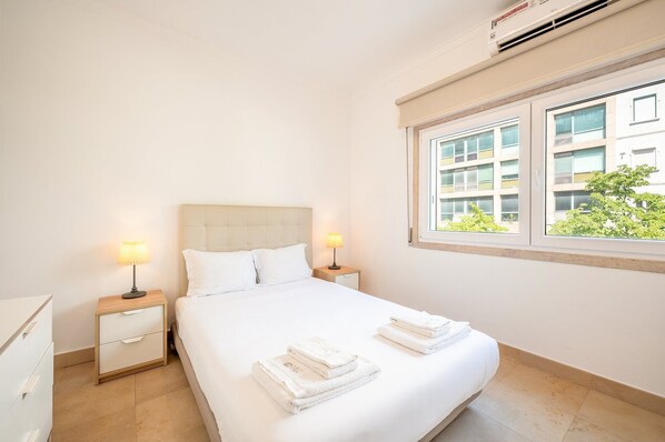A hotel room with a bed, two bedside tables with lamps, a window with a view of a building and greenery, and a ceiling air conditioner. - GuestReady - Almirante Avenue (Lisbon)