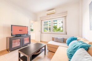 Una sala de estar moderna con televisor de pantalla plana, un sofá con cojines, una mesa de centro y una ventana con vistas a edificios.