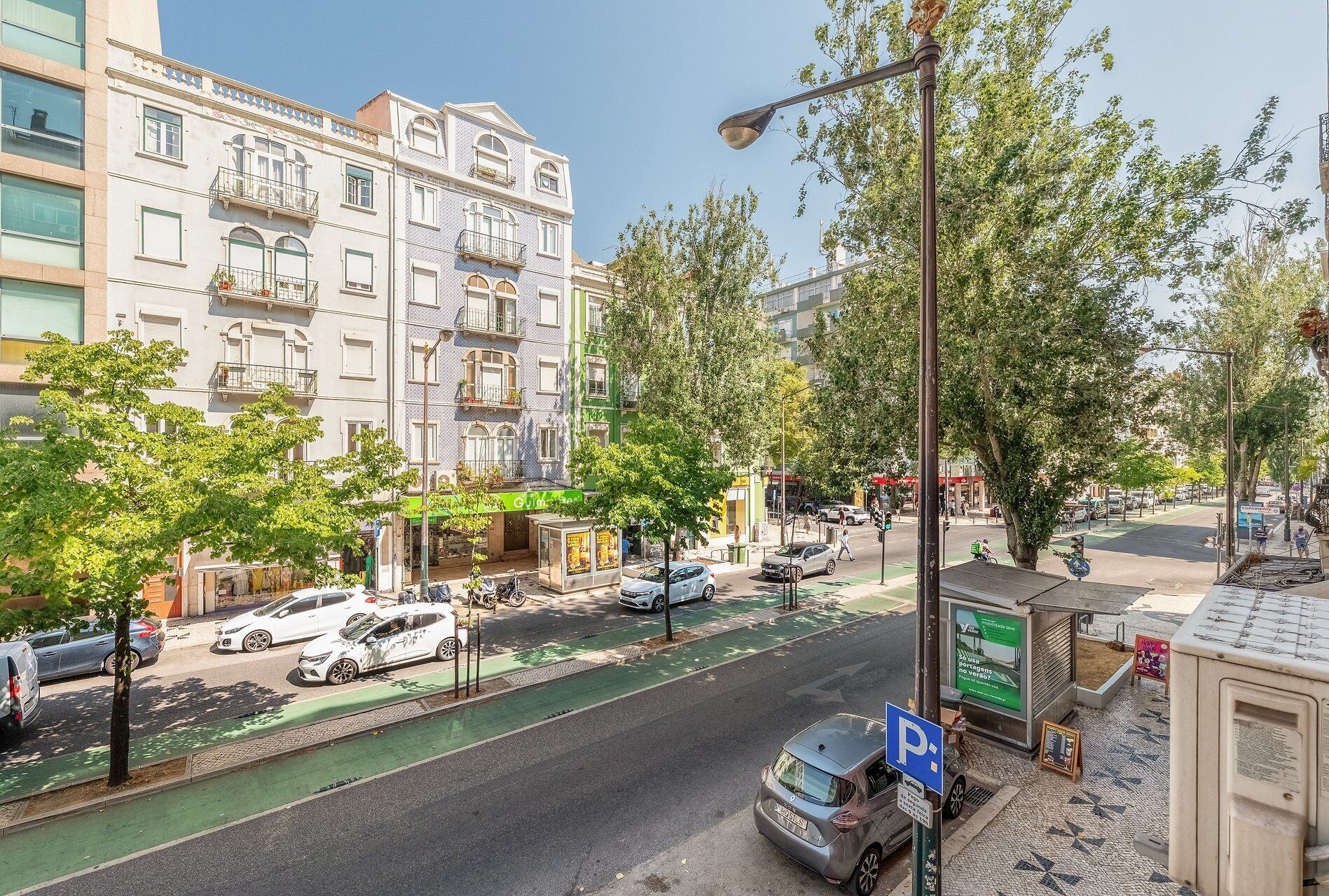 Vista da rua com carros estacionados, ponto de ônibus e um edifício com vários andares e sacadas.
