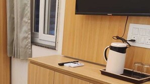 A compact room with a wooden desk, a television mounted on the wall, a window with curtains, and a kettle on the desk. - Laxmi Seva Sadan Laxmi Hotel  (Barsana)