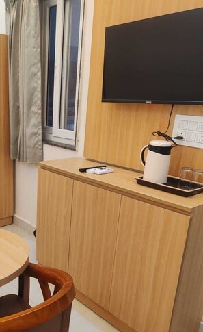 A compact room with a wooden desk, a television mounted on the wall, a window with curtains, and a kettle on the desk. - Laxmi Seva Sadan Laxmi Hotel  (Barsana)