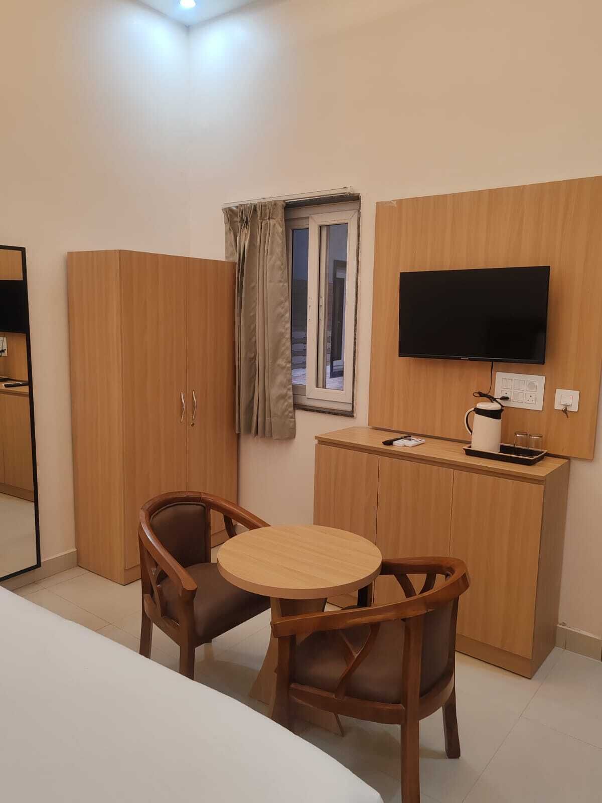 Ein Hotelzimmer mit Bett, Fernseher, Holzschrank, kleinem Tisch mit Stühlen und einem Fenster mit Vorhängen.