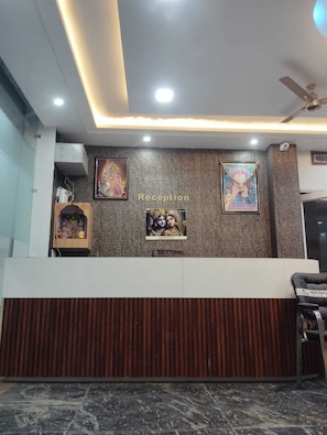 A reception area with a modern design, featuring a wooden panel wall, a ceiling fan, and framed religious posters. - Laxmi Seva Sadan Laxmi Hotel  (Barsana)