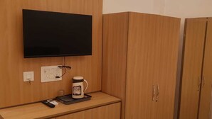 A room with a flat-screen TV mounted on a wooden wall, a small table with a coffee maker, and a bed with a white mattress. - Laxmi Seva Sadan Laxmi Hotel  (Barsana)