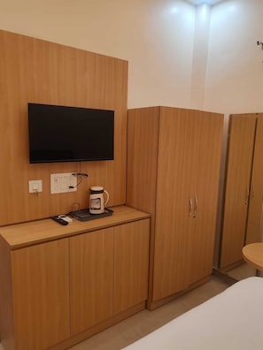 A room with a flat-screen TV mounted on a wooden wall, a small table with a coffee maker, and a bed with a white mattress. - Laxmi Seva Sadan Laxmi Hotel  (Barsana)