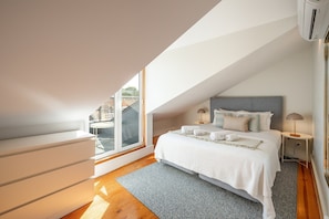 Ein Schlafzimmer mit großem Fenster, einem Bett mit Kissen, einem Nachttisch und einem Teppich.