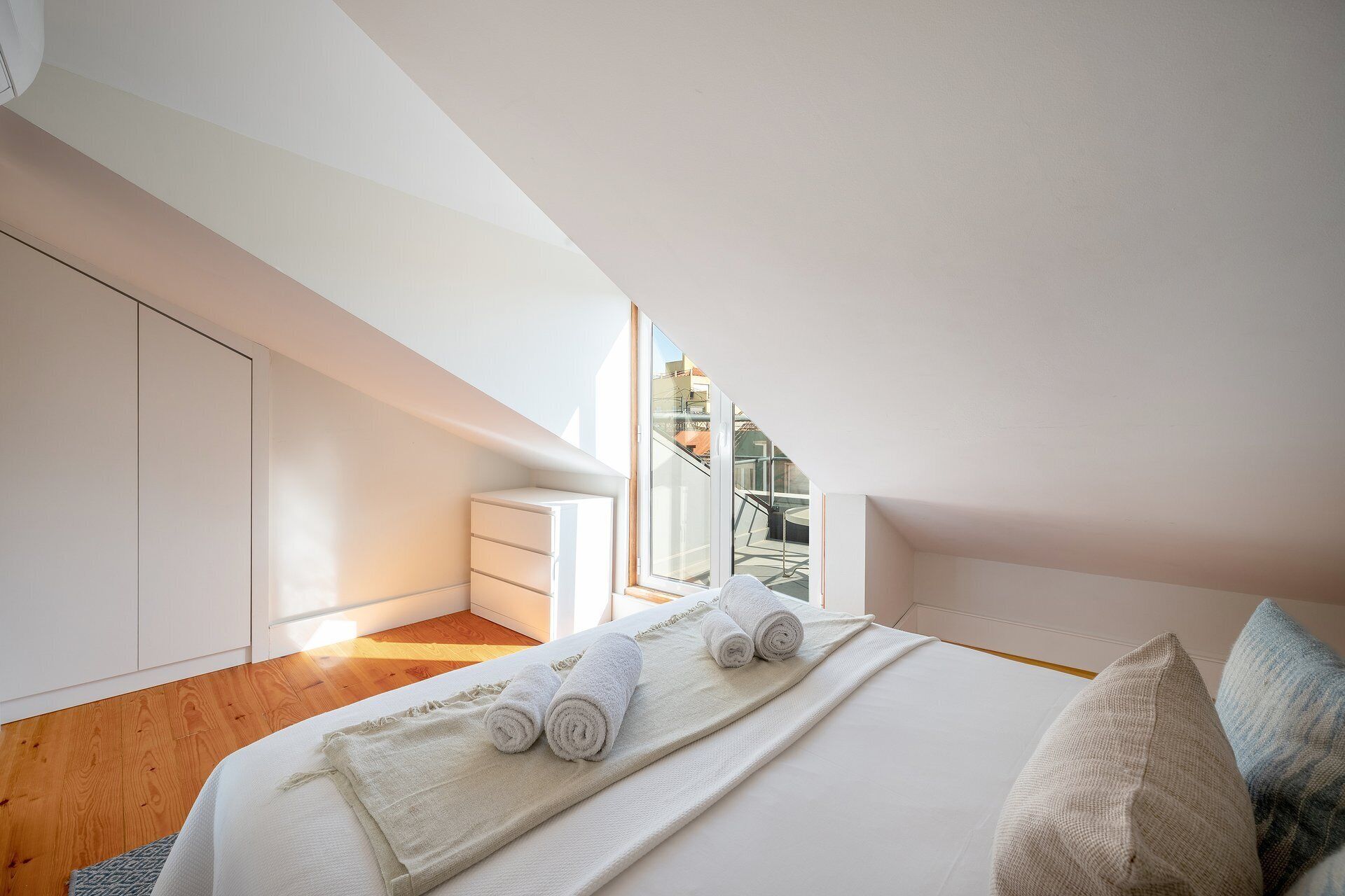 Un dormitorio con tragaluz, una cama con toallas enrolladas, una mesita de noche y vistas de la ciudad a través de la ventana.