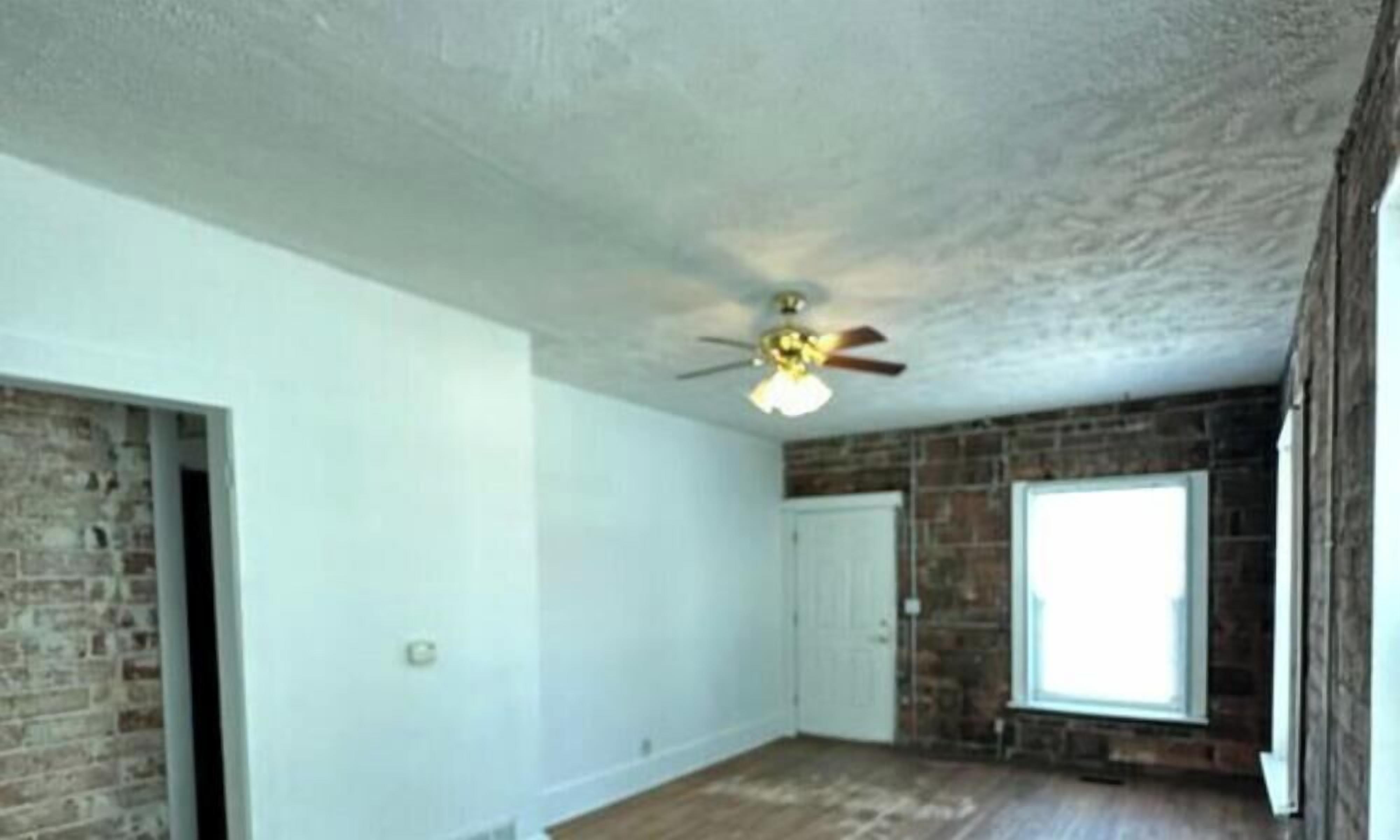 Une pièce avec un ventilateur de plafond, un mur en briques, un plancher en bois et une fenêtre.