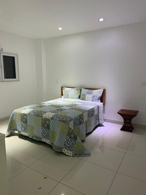 A bedroom with a bed, a wooden nightstand, and a window with blinds. - Holiday Homes - Newtown Apartment 1 (Nadi)