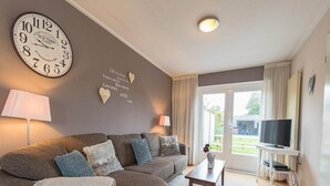 A living room with a sofa, a coffee table, a TV, a clock, and a wall with heart-shaped decorations. - Holiday Home With Private Parking (Oostkapelle)