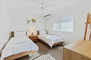 Une chambre avec deux lits, un ventilateur de plafond, un appareil de climatisation, une fenêtre avec des stores, une commode en bois et une petite table.