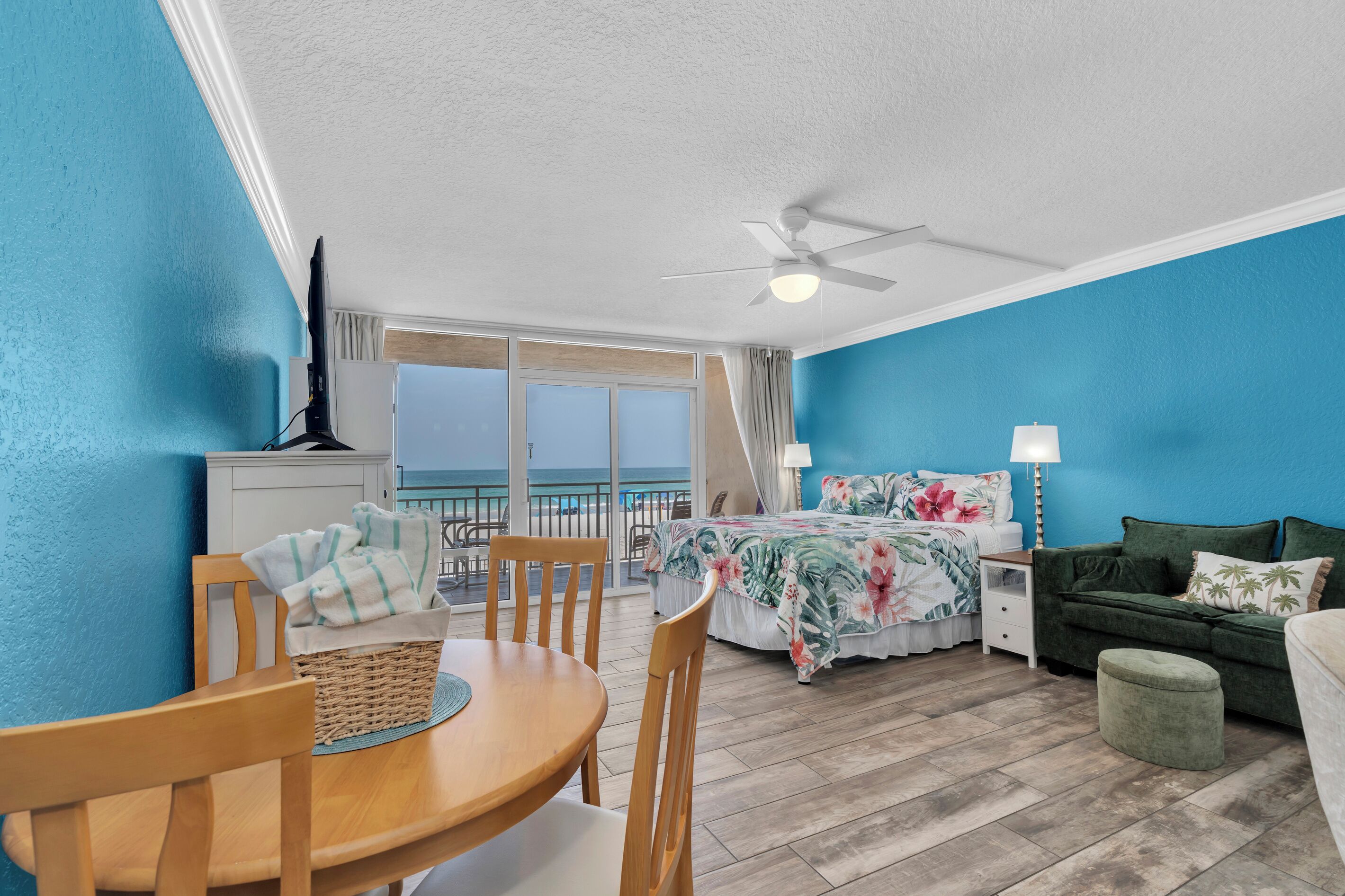 A room with a bed, a table with chairs, a ceiling fan, and a view of the ocean.