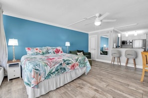 A bedroom with a bed, two bedside lamps, a ceiling fan, and a view into the adjacent kitchen area. - Beachfront Studio on Anna Maria Island (Bradenton Beach)