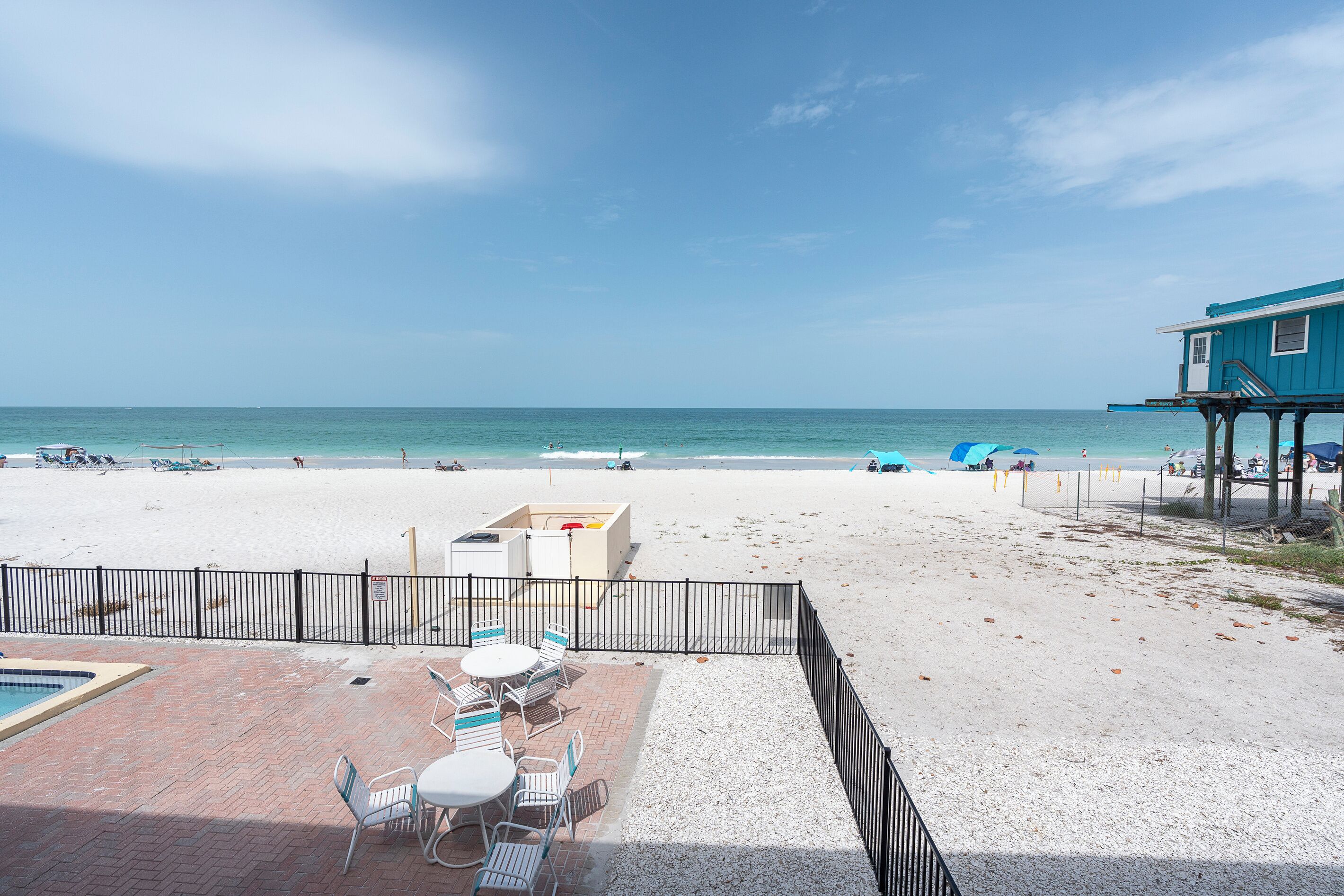 A beach view with a pool, patio chairs, and a building with a balcony.