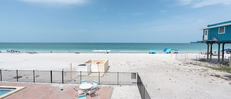 A beach view with a pool, patio chairs, and a building with a balcony.