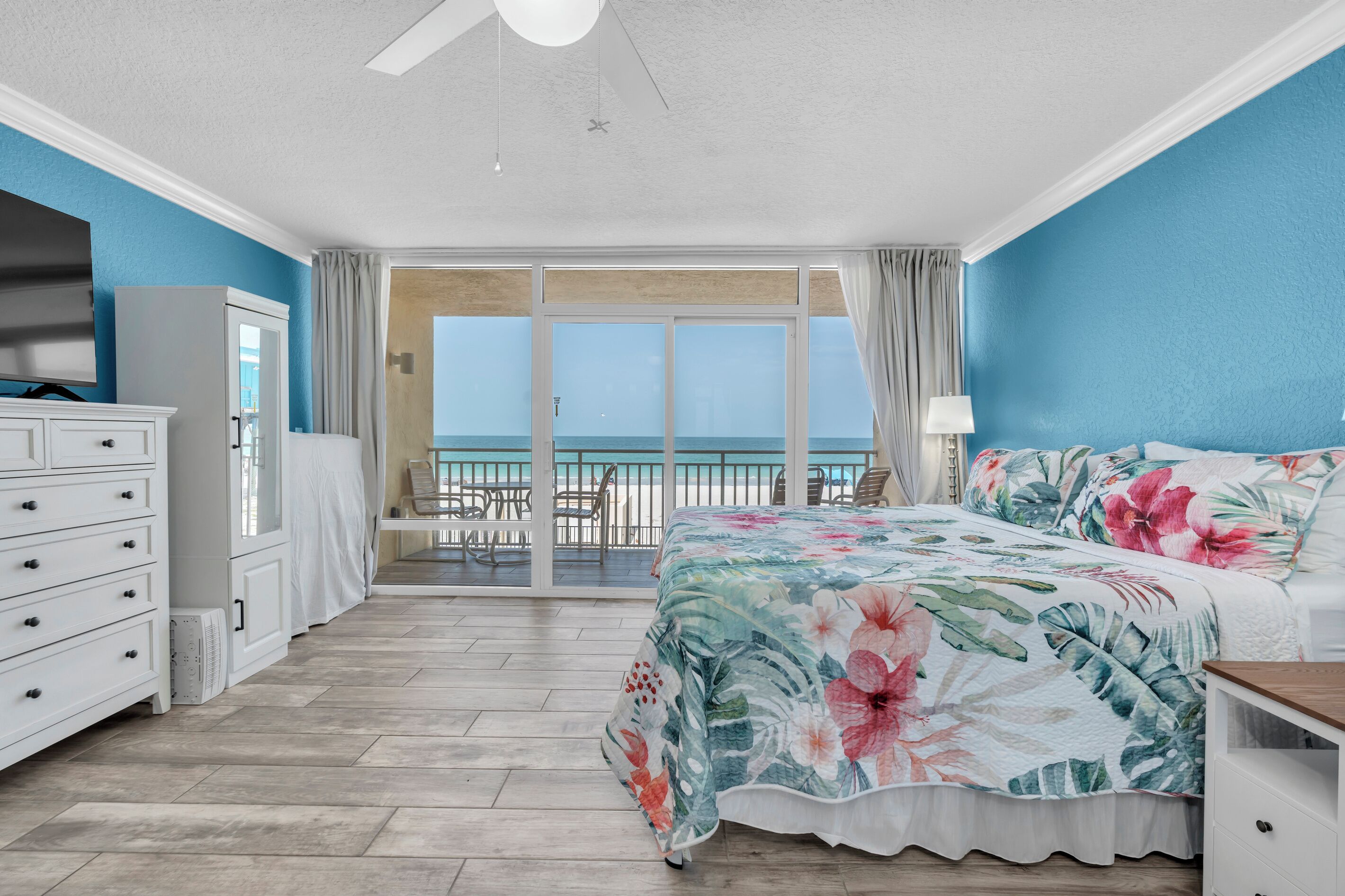 A bedroom with a large bed, a dresser, a ceiling fan, and a view of the ocean through a sliding glass door.