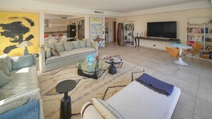 En moderne stue med sofa, sofabord, fjernsyn, bogreol og trappe til køkkenet.