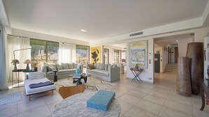 En rummelig stue med sofa, sofabord og spiseplads. Der er et stort vindue der giver naturligt lys og udsigt til det fri.