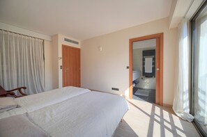 Ein Schlafzimmer mit einem Bett, einer Holztür, einem durch eine offene Tür einsehbaren Badezimmer und einem mit Vorhängen drapierten Fenster.