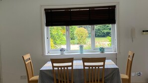 A dining area with a table and four chairs, a window with blinds, and a view of greenery outside. - The Bungalow offers 2 private, spacious and stylish ensuite bedrooms (West End, Woking)