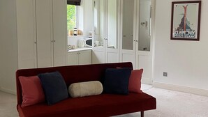 A red sofa with cushions, white wardrobes, a kitchen area, and a framed picture on the wall. - The Bungalow offers 2 private, spacious and stylish ensuite bedrooms (West End, Woking)