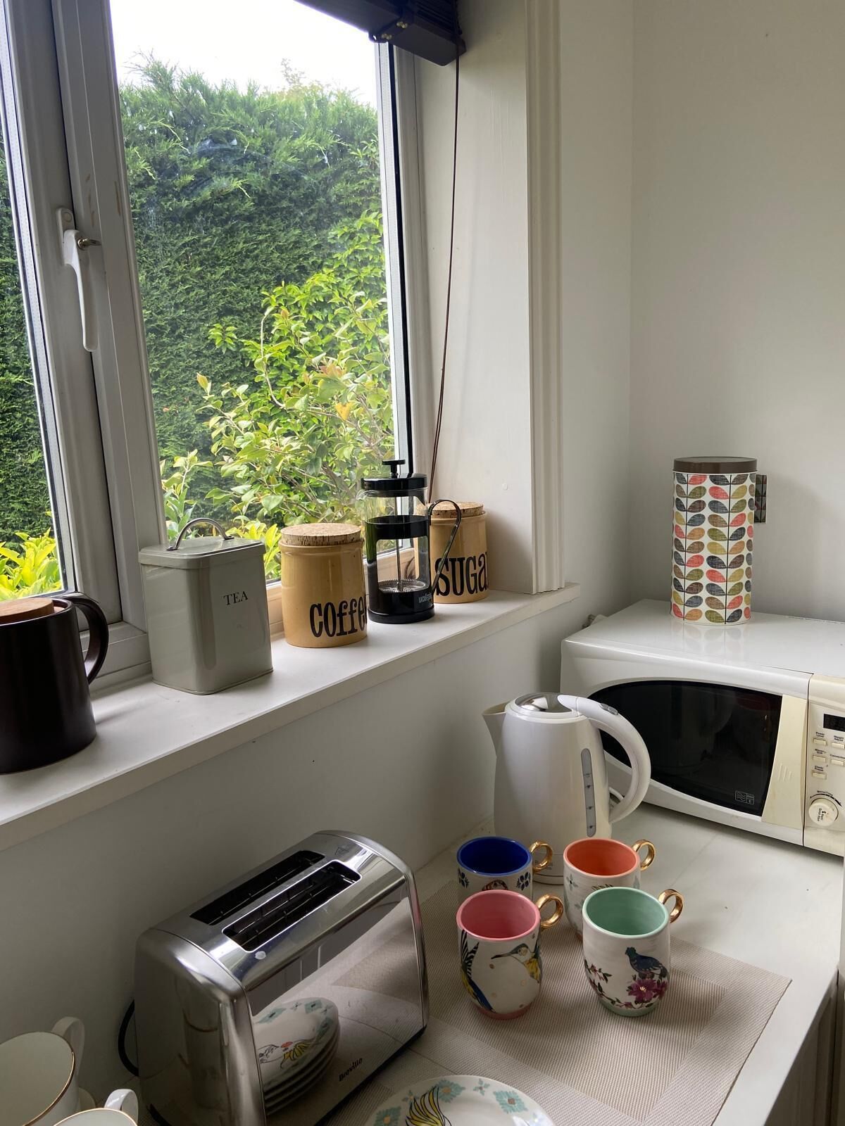 A kitchen with a toaster, a kettle, and a microwave on a white countertop. There are three mugs with floral designs and a sugar container on the windowsill.