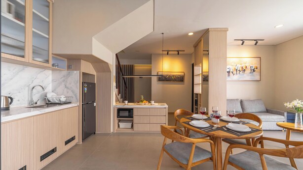 Una cocina moderna con refrigerador incorporado, una zona de comedor con sillas de madera y una mesa dispuesta para una comida, y una sala de estar con sofá y un cuadro en la pared.