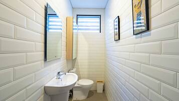 Un baño estrecho con paredes de azulejos blancos, un lavamanos blanco, un inodoro y una ventana con persianas.