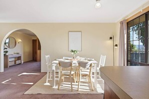 A dining area with a round table, white chairs, and a rug. There is a window with curtains, a door leading to another room, and a wall-mounted light fixture.