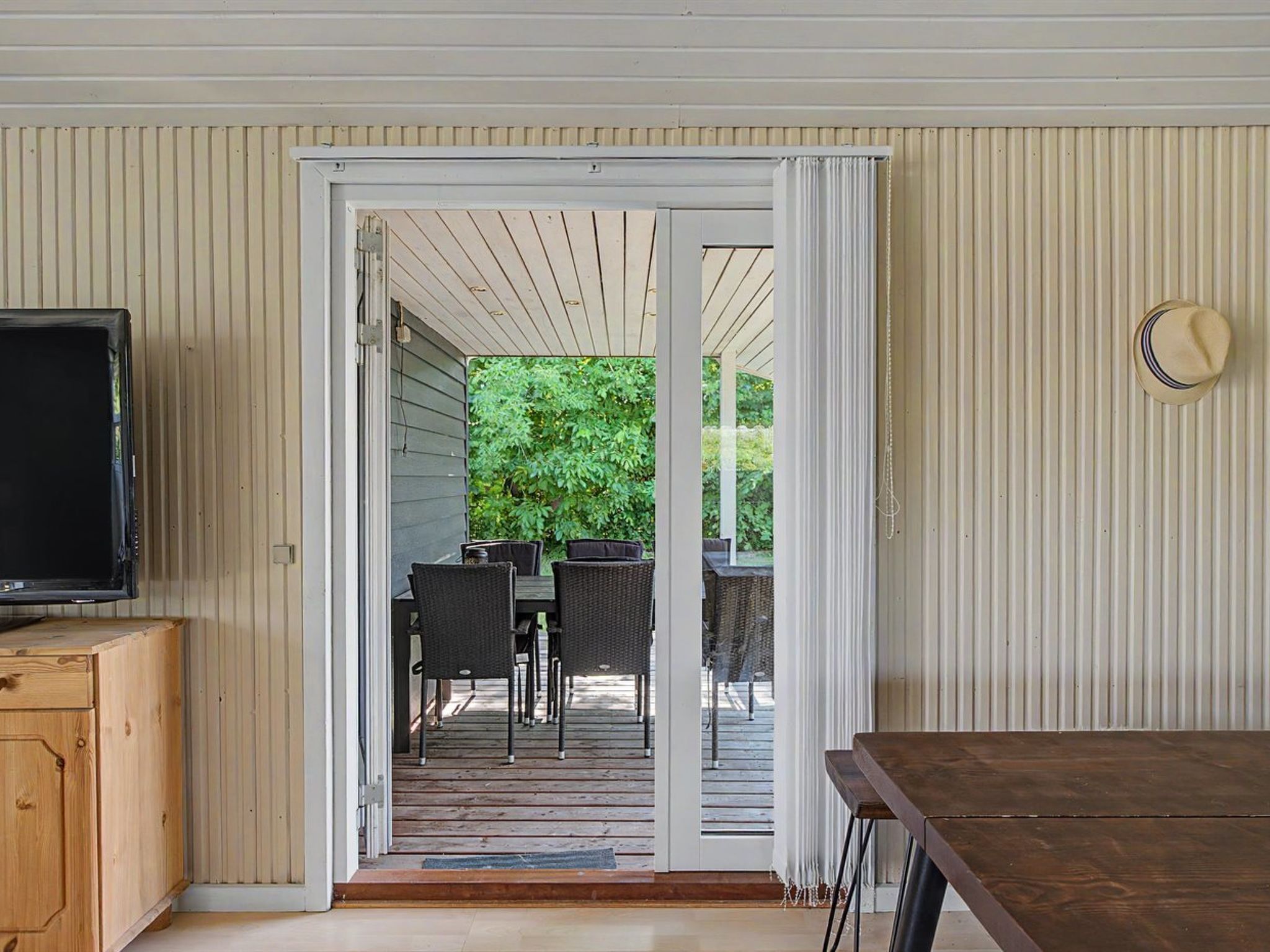 Een kamer met een televisie op een houten kast, een glazen schuifdeur naar een terras buiten met een tafel en stoelen.