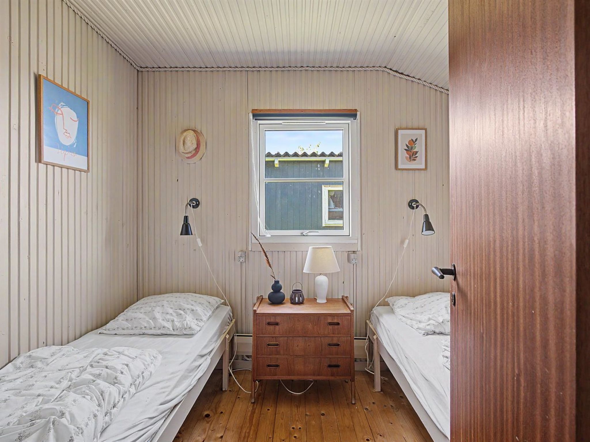 A bedroom with two single beds, a wooden dresser, a window with a view, and two wall-mounted lamps.