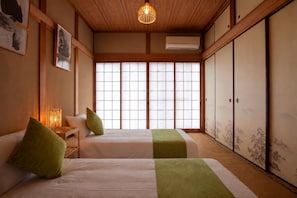 Una habitación tradicional japonesa con suelo de tatami, dos camas con detalles verdes, una ventana con puertas correderas shoji y una lámpara colgante.