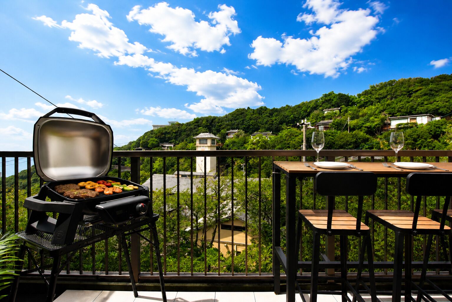 A balcony with a grill, dining table, chairs, and a view of a lush green landscape.