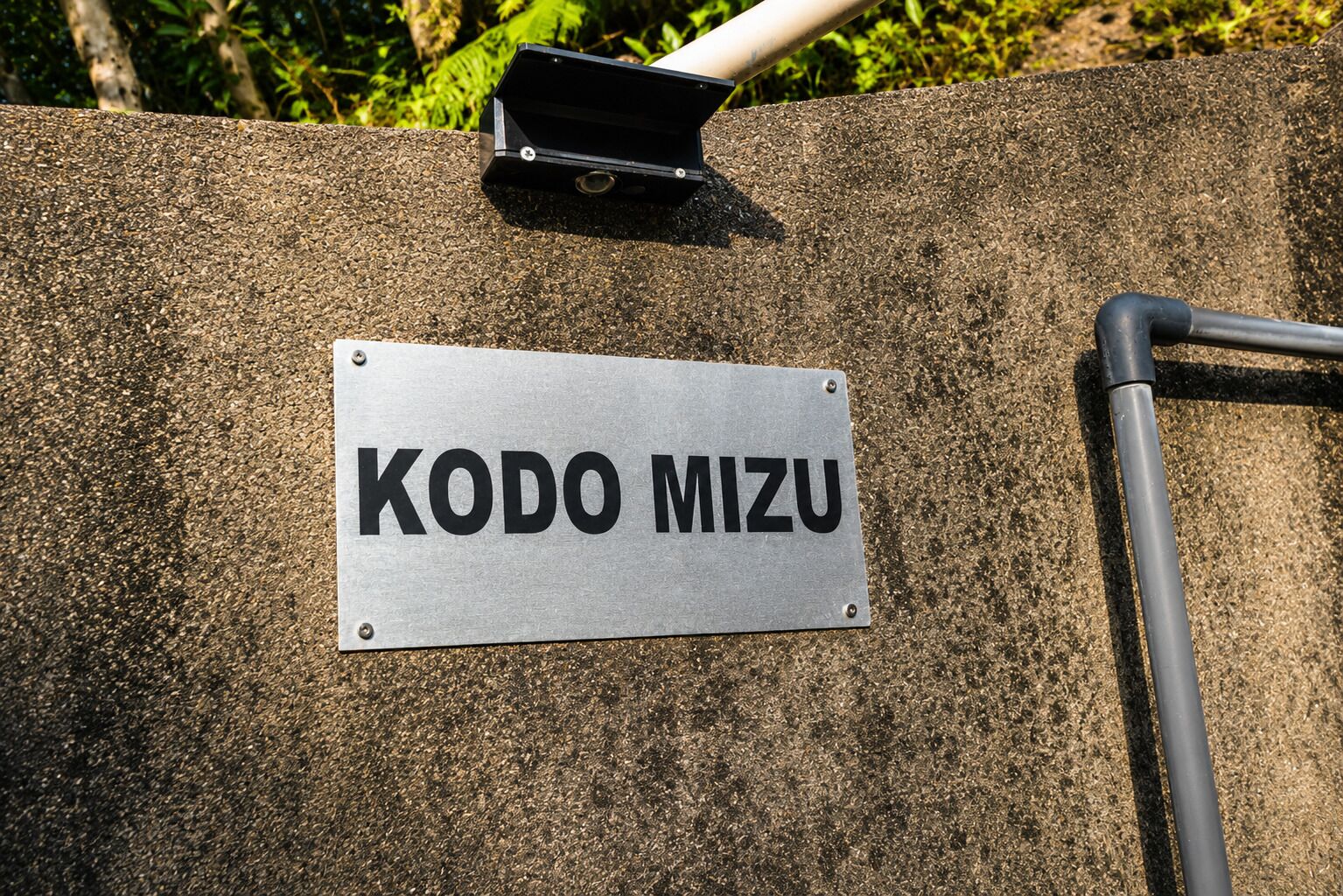 Un letrero metálico con el texto 'KODO MIZU' montado sobre una superficie texturizada, presumiblemente una pared, con una estructura metálica encima.