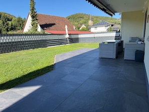 A covered patio area with a tiled floor, outdoor furniture, and a view of a hillside with buildings.
